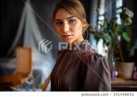 An young woman in vintage long dress standing in moody interior with studio props and natural window An young woman in vintage long dress standing in moody interior with studio props and natural window 141436658