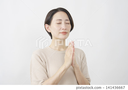 A woman in her 40s with her eyes closed and hands clasped in prayer against a white background. A woman in her 40s with her eyes closed and hands clasped in prayer against a white background. 141436667