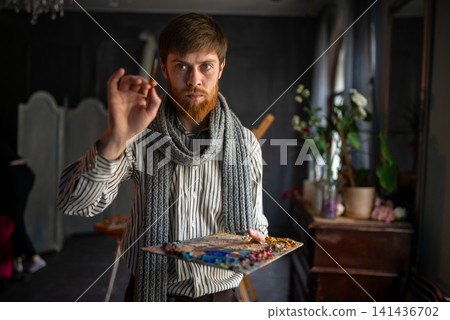 Bearded artist mixing oil paint on wooden palette in dim studio with plants and vintage furniture 141436702