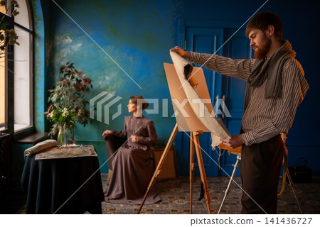Male artist painting portrait of seated woman in vintage studio with teal wall and window light Male artist painting portrait of seated woman in vintage studio with teal wall and window light 141436727
