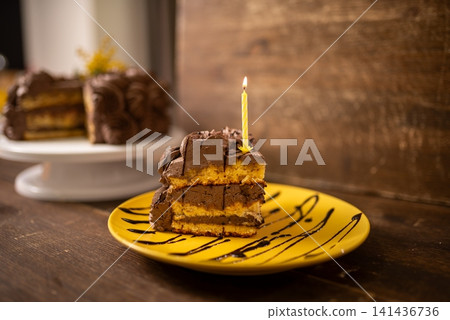 Slice of layered chocolate cake with lit candle on yellow plate against rustic wood background Slice of layered chocolate cake with lit candle on yellow plate against rustic wood background 141436736