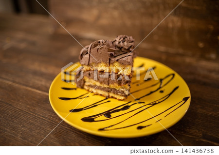 Chocolate layer cake slice on yellow plate with chocolate drizzle in warm cafe lighting Chocolate layer cake slice on yellow plate with chocolate drizzle in warm cafe lighting 141436738