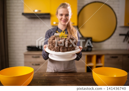 Smiling woman presenting chocolate cake with decorative topping in modern kitchen setting Smiling woman presenting chocolate cake with decorative topping in modern kitchen setting 141436740