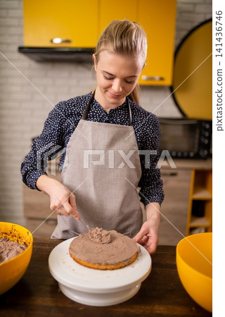 Smiling woman in apron frosting round chocolate cake on turntable in warm modern kitchen Smiling woman in apron frosting round chocolate cake on turntable in warm modern kitchen 141436746