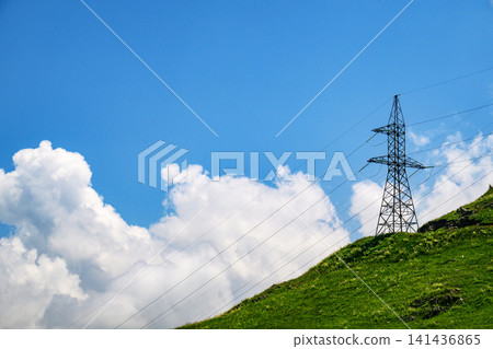 Electrical transmission tower on grassy hill against bright blue sky and clouds Electrical transmission tower on grassy hill against bright blue sky and clouds 141436865