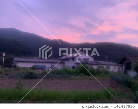 山村景色，房屋和田野依偎在山脈中，映襯著泛著紫色光暈的夕陽天空。 141437050