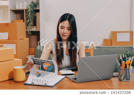Startup SME small business entrepreneur of freelance Asian woman wearing apron using laptop and box to receive and review orders online to prepare to pack sell to customers, online sme business ideas. Startup SME small business entrepreneur of freelance Asian woman wearing apron using laptop and box to receive and review orders online to prepare to pack sell to customers, online sme business ideas. 141437206