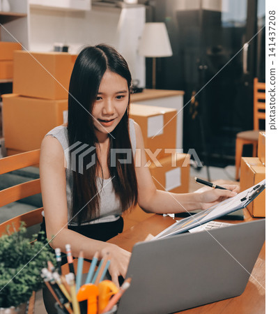 Startup SME small business entrepreneur of freelance Asian woman wearing apron using laptop and box to receive and review orders online to prepare to pack sell to customers, online sme business ideas. Startup SME small business entrepreneur of freelance Asian woman wearing apron using laptop and box to receive and review orders online to prepare to pack sell to customers, online sme business ideas. 141437208