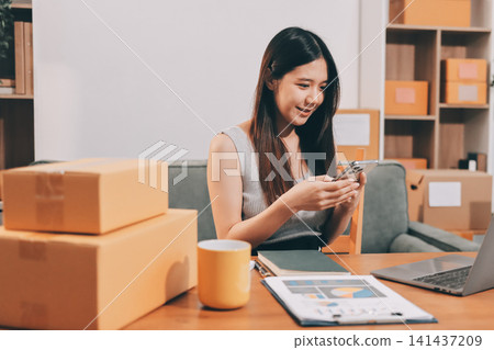 Startup SME small business entrepreneur of freelance Asian woman wearing apron using laptop and box to receive and review orders online to prepare to pack sell to customers, online sme business ideas. Startup SME small business entrepreneur of freelance Asian woman wearing apron using laptop and box to receive and review orders online to prepare to pack sell to customers, online sme business ideas. 141437209