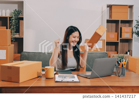 Startup SME small business entrepreneur of freelance Asian woman wearing apron using laptop and box to receive and review orders online to prepare to pack sell to customers, online sme business ideas. Startup SME small business entrepreneur of freelance Asian woman wearing apron using laptop and box to receive and review orders online to prepare to pack sell to customers, online sme business ideas. 141437217