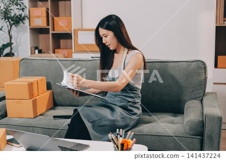 Startup SME small business entrepreneur of freelance Asian woman wearing apron using laptop and box to receive and review orders online to prepare to pack sell to customers, online sme business ideas. Startup SME small business entrepreneur of freelance Asian woman wearing apron using laptop and box to receive and review orders online to prepare to pack sell to customers, online sme business ideas. 141437224