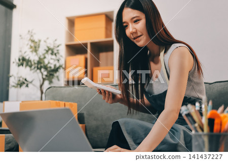 Startup SME small business entrepreneur of freelance Asian woman wearing apron using laptop and box to receive and review orders online to prepare to pack sell to customers, online sme business ideas. Startup SME small business entrepreneur of freelance Asian woman wearing apron using laptop and box to receive and review orders online to prepare to pack sell to customers, online sme business ideas. 141437227