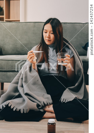 sick asian young woman sneezing into tissue paper while covered with a blanket and sitting on sofa in the living room sick asian young woman sneezing into tissue paper while covered with a blanket and sitting on sofa in the living room 141437254
