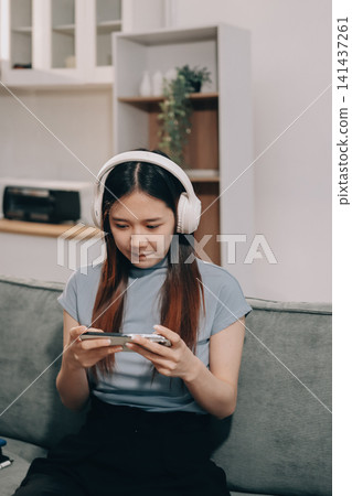 Beautiful young asian woman sitting on sofa and holding joystick playing online video game in living room at house Beautiful young asian woman sitting on sofa and holding joystick playing online video game in living room at house 141437261