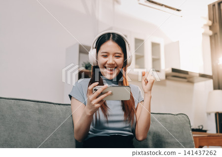 Beautiful young asian woman sitting on sofa and holding joystick playing online video game in living room at house Beautiful young asian woman sitting on sofa and holding joystick playing online video game in living room at house 141437262