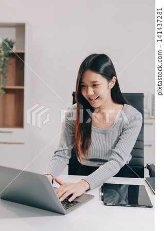 Young asian businesswoman is smiling and working on a laptop while sitting at her desk in a modern office Young asian businesswoman is smiling and working on a laptop while sitting at her desk in a modern office 141437281