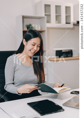 Young asian businesswoman is smiling and working on a laptop while sitting at her desk in a modern office Young asian businesswoman is smiling and working on a laptop while sitting at her desk in a modern office 141437282