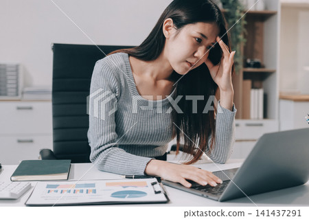 Young Asian woman sits at office desk rubbings her eyes and temples feeling pain. Stress. And exhaustion from long hours working on the computer 141437291