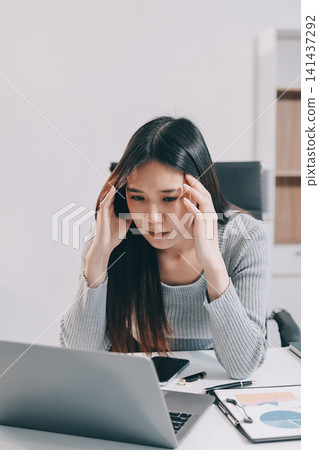 Young Asian woman sits at office desk rubbings her eyes and temples feeling pain. Stress. And exhaustion from long hours working on the computer Young Asian woman sits at office desk rubbings her eyes and temples feeling pain. Stress. And exhaustion from long hours working on the computer 141437292