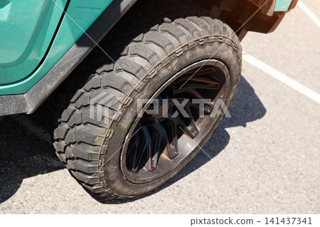Close-up detail of rugged off-road tire on green vehicle fender, showcasing custom black wheel with red accents, strong tread pattern, parked outdoors on asphalt during bright day Close-up detail of rugged off-road tire on green vehicle fender, showcasing custom black wheel with red accents, strong tread pattern, parked outdoors on asphalt during bright day 141437341