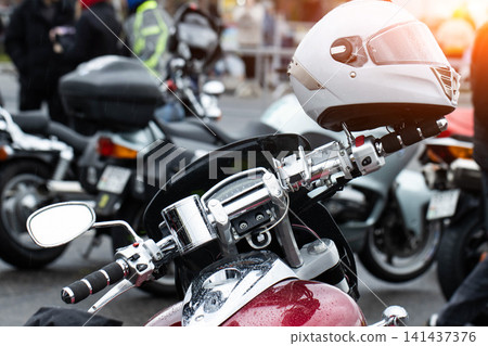 Modern motorcycle with white helmet on handlebars, covered in raindrops, parked outdoors during day, chrome details reflecting light, street scene with other vehicles and blurred people background. 141437376