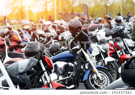 Row of parked motorcycles with helmets resting on seats, showcasing diverse collection of bikes at outdoor gathering during bright daytime, lens flare adding striking luminous effect to overall scene. Row of parked motorcycles with helmets resting on seats, showcasing diverse collection of bikes at outdoor gathering during bright daytime, lens flare adding striking luminous effect to overall scene. 141437381