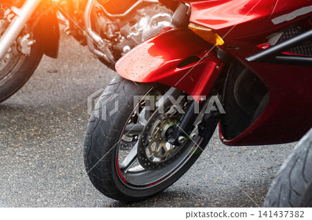 Close up view of front wheel, tire, and red fender of sport motorcycle with water drops on wet asphalt ground, blurred other motorcycles in background with warm light lens flare. Close up view of front wheel, tire, and red fender of sport motorcycle with water drops on wet asphalt ground, blurred other motorcycles in background with warm light lens flare. 141437382