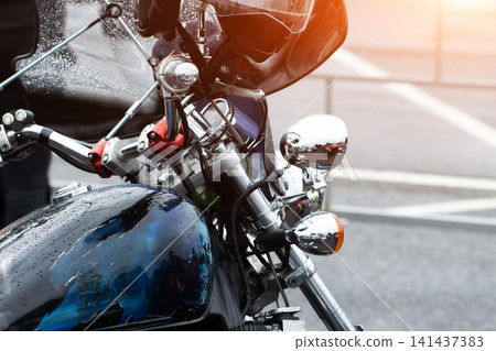 Close-up detail of motorcycle front section with handlebars, fuel tank, chrome elements, and rearview mirror showing raindrops, sun flare on windshield, custom paint artwork on tank, street background 141437383