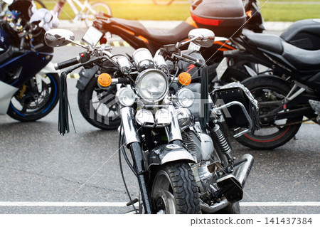 Close up front view of a black cruiser motorcycle with chrome details and leather tassel on handlebar, parked on wet asphalt street with other motorcycles in background on a cloudy day. Close up front view of a black cruiser motorcycle with chrome details and leather tassel on handlebar, parked on wet asphalt street with other motorcycles in background on a cloudy day. 141437384