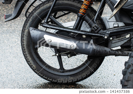 Motorcycle rear wheel and exhaust system close-up on wet asphalt surface. Details of suspension, tire tread, and metal components after rain, showcasing transportation vehicle parts and mechanics. Motorcycle rear wheel and exhaust system close-up on wet asphalt surface. Details of suspension, tire tread, and metal components after rain, showcasing transportation vehicle parts and mechanics. 141437386