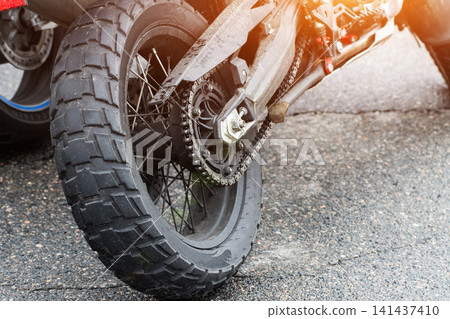 Close up view of motorcycle rear wheel and drive chain with knobby tire tread on paved road detail of vehicle engineering and power transmission system with sunlight Close up view of motorcycle rear wheel and drive chain with knobby tire tread on paved road detail of vehicle engineering and power transmission system with sunlight 141437410