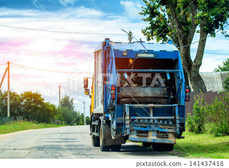 Blue garbage truck back view parked on cracked asphalt road during daytime. Waste collection vehicle working on rural street with green trees and cloudy sky. Copy space for text Blue garbage truck back view parked on cracked asphalt road during daytime. Waste collection vehicle working on rural street with green trees and cloudy sky. Copy space for text 141437418