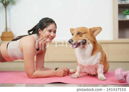 Asian woman doing plank exercise on yoga mat looking at her corgi dog Asian woman doing plank exercise on yoga mat looking at her corgi dog 141437550