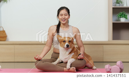 Asian woman meditating in lotus pose on yoga mat with corgi dog Asian woman meditating in lotus pose on yoga mat with corgi dog 141437551