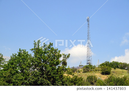 Telecom tower rising over grassy hill with trees below 141437803