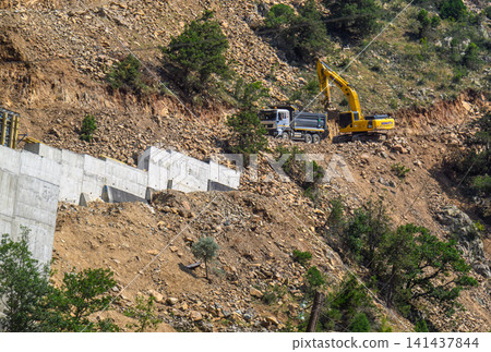 Excavator works on steep, dry hillside with concrete wall Excavator works on steep, dry hillside with concrete wall 141437844