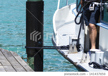 Man securing a boat with mooring ropes at a pier over turquoise water, illustrating maritime work and dockside responsibilities. Man securing a boat with mooring ropes at a pier over turquoise water, illustrating maritime work and dockside responsibilities. 141437850