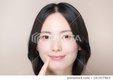 一位笑容甜美、嘴唇豐滿的日本女子在粉米色的背景下直視鏡頭,並指著自己的嘴唇。 一位笑容甜美、嘴唇豐滿的日本女子在粉米色的背景下直視鏡頭,並指著自己的嘴唇。 141437863
