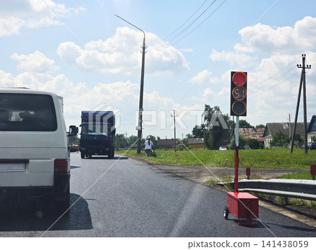 White van and dark truck stopped on newly paved asphalt road at portable red traffic light showing countdown in rural village area on sunny day with blue sky and white clouds, reversible traffic White van and dark truck stopped on newly paved asphalt road at portable red traffic light showing countdown in rural village area on sunny day with blue sky and white clouds, reversible traffic 141438059