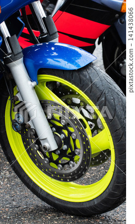 Close-up of front wheel of motorcycle with vibrant neon yellow rim on wet asphalt after rain. Blue fender, silver forks, red body panel visible, showcasing detail of braking system and tire tread. Close-up of front wheel of motorcycle with vibrant neon yellow rim on wet asphalt after rain. Blue fender, silver forks, red body panel visible, showcasing detail of braking system and tire tread. 141438066