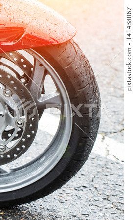 Close up view of front wheel of red motorcycle on wet asphalt road, fender covered in raindrops, detail of tire, rim, and brake discs, vertical composition showing vehicle part during rainy weather Close up view of front wheel of red motorcycle on wet asphalt road, fender covered in raindrops, detail of tire, rim, and brake discs, vertical composition showing vehicle part during rainy weather 141438067