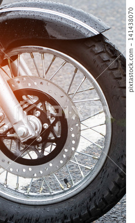 Close up detail of motorcycle front wheel, wet tire, chrome spokes, and disc brake after rain. Wet road surface. Vertical composition. Close up detail of motorcycle front wheel, wet tire, chrome spokes, and disc brake after rain. Wet road surface. Vertical composition. 141438070