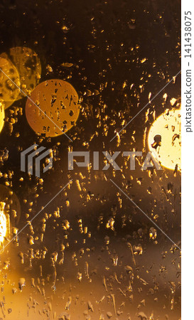 Water droplets on glass surface creating abstract bokeh effect from night city lights, golden yellow reflections in dark urban environment, rainy weather atmosphere vertical photo Water droplets on glass surface creating abstract bokeh effect from night city lights, golden yellow reflections in dark urban environment, rainy weather atmosphere vertical photo 141438075