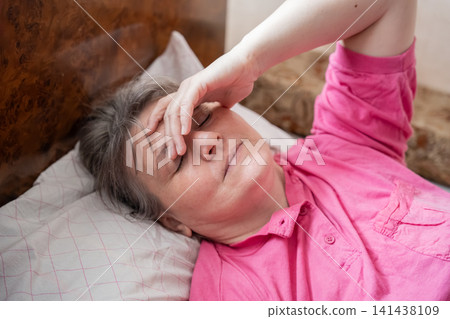Middle-aged woman lying in bed with hand on forehead expressing discomfort. Menopause fatigue headache management and sleep disturbance Middle-aged woman lying in bed with hand on forehead expressing discomfort. Menopause fatigue headache management and sleep disturbance 141438109