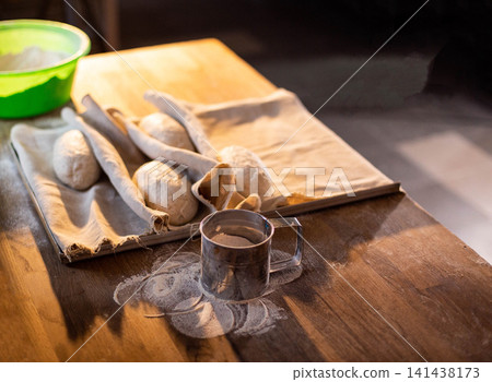 Freshly kneaded bread dough rounds resting on linen cloth in proofing basket, preparation for baking on wooden kitchen table with flour dusting and sifter, homemade bakery goods production Freshly kneaded bread dough rounds resting on linen cloth in proofing basket, preparation for baking on wooden kitchen table with flour dusting and sifter, homemade bakery goods production 141438173