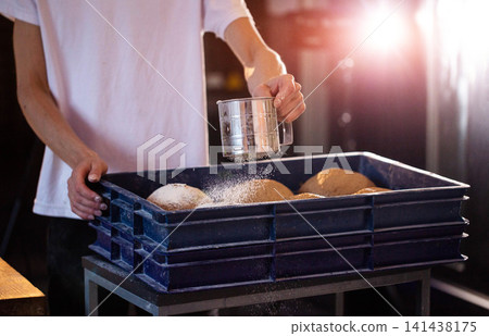 Baker hands sift flour over bread dough in blue crate manual preparation of artisan bakery products at factory bread production facility bright light Baker hands sift flour over bread dough in blue crate manual preparation of artisan bakery products at factory bread production facility bright light 141438175