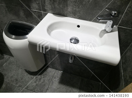 Modern bathroom interior features white ceramic sink with chrome faucet and drain, alongside sleek white and grey hand dryer waste bin unit on dark grey tiled walls and floor Modern bathroom interior features white ceramic sink with chrome faucet and drain, alongside sleek white and grey hand dryer waste bin unit on dark grey tiled walls and floor 141438193