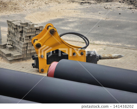 Yellow hydraulic hammer attachment with sharp metal point rests on ground at construction site, alongside stack of grey paving stones and large black industrial pipes awaiting installation Yellow hydraulic hammer attachment with sharp metal point rests on ground at construction site, alongside stack of grey paving stones and large black industrial pipes awaiting installation 141438202