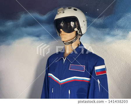 Mannequin display of pilot flight suit and helmet with visor, featuring blue jumpsuit, white helmet, and chest stripes against painted wall background, showcasing aviation attire. Mannequin display of pilot flight suit and helmet with visor, featuring blue jumpsuit, white helmet, and chest stripes against painted wall background, showcasing aviation attire. 141438203