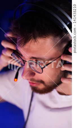 Young man in glasses and headphones with illuminated microphone adjusts headset, concentrated expression during online gaming or communication session, blue background light, vertical photo 141438210